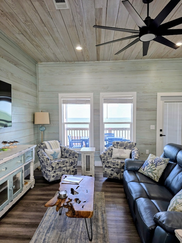 A living room with a wooden ceiling, a ceiling fan, two windows with a view, a TV, a coffee table, a sofa, and a side table. - Dancing Dolphin-Waterfront! Hot Tub, Arcade, Pool, Gas Grill-Walk to Pier & Pubs (Fulton)
