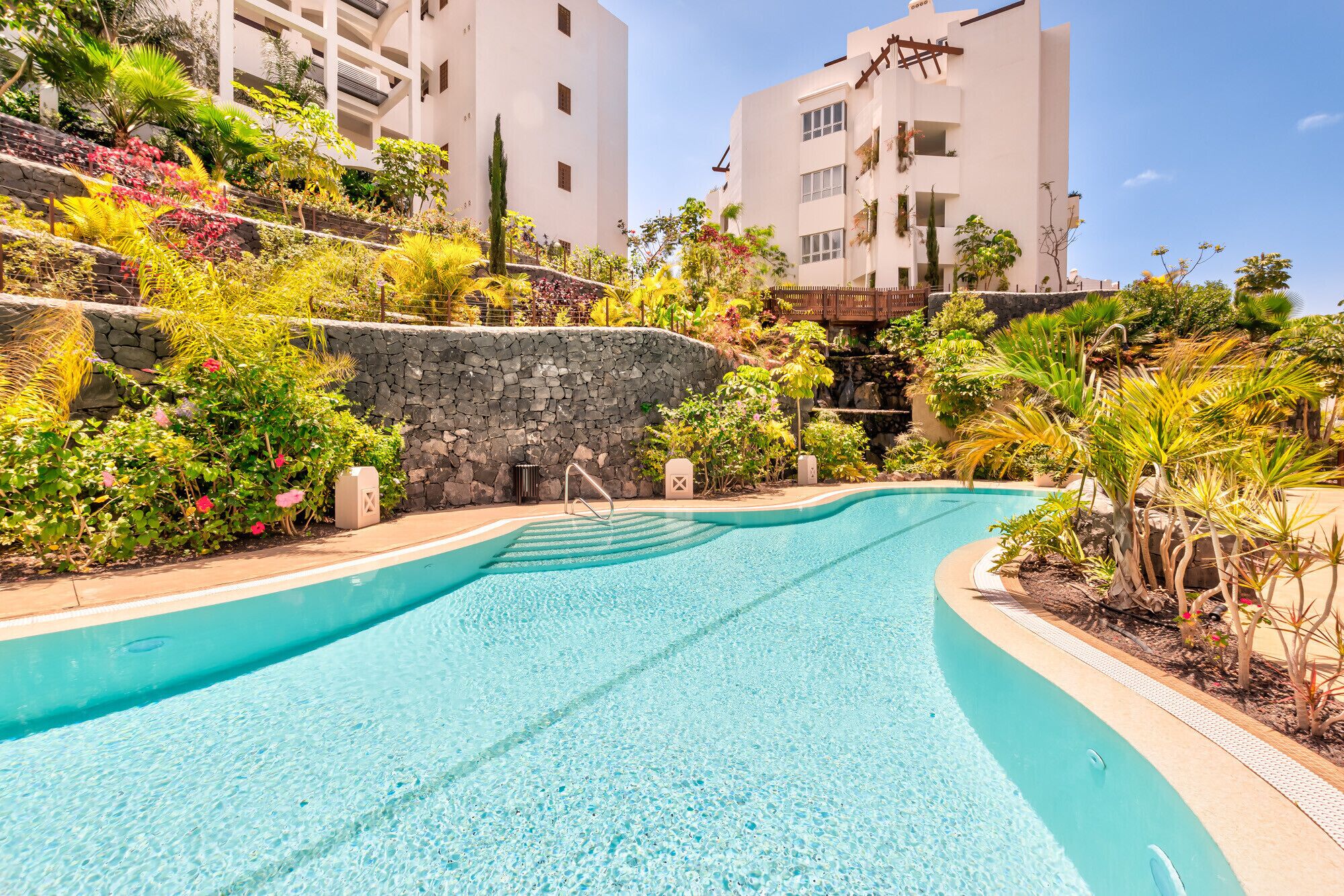 A swimming pool surrounded by lush greenery and stone walls, with modern buildings in the background.