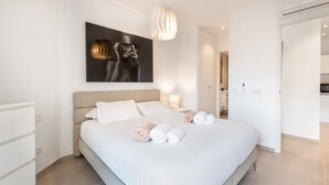 A modern hotel bedroom with a large bed, a bedside table with a lamp, a wall-mounted artwork, and an air conditioning unit.