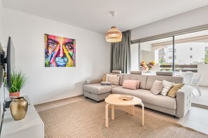 A modern living room with a sofa, a small round table, a hanging light fixture, a large window, and a painting on the wall.