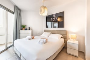 A modern hotel room with a large bed, a bedside table with a lamp, a wall-mounted artwork, and a view of the outdoors.
