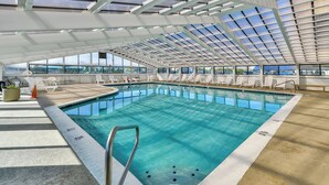 Piscina interna com água azul cristalina, rodeada por espreguiçadeiras e teto de vidro.