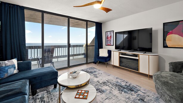Uma sala de estar moderna com uma televisão grande, um ventilador de teto e vista para o mar.