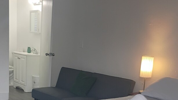 Ein Schlafzimmer mit Bett, Nachttischlampe, einem Badezimmer inklusive Waschbecken und Toilette sowie einer Tür zu einem weiteren Raum.