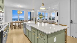 A modern kitchen with a large island, pendant lighting, and a view of the ocean. - Oceanfront Luxury | 4BR w/ Elevator sleeps 12 (North Topsail Beach)
