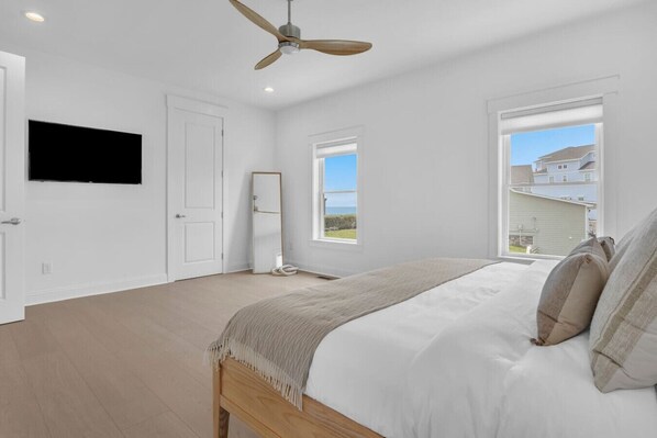 A bedroom with a bed, a ceiling fan, a TV, and a window with a view of a house and sky.