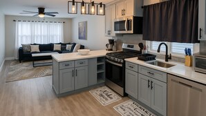 Een moderne keuken met grijze kastjes, een zwarte kookplaat en een magnetron. Er is een zithoek met een donkergrijze bank en een koffiezetapparaat.