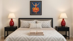 Een net opgemaakt bed met een geometrisch beddegoed, twee nachtkastjes met rode lampen, een ingelijst beeld boven het bed, en een bijpassende bank aan het voeteneinde.