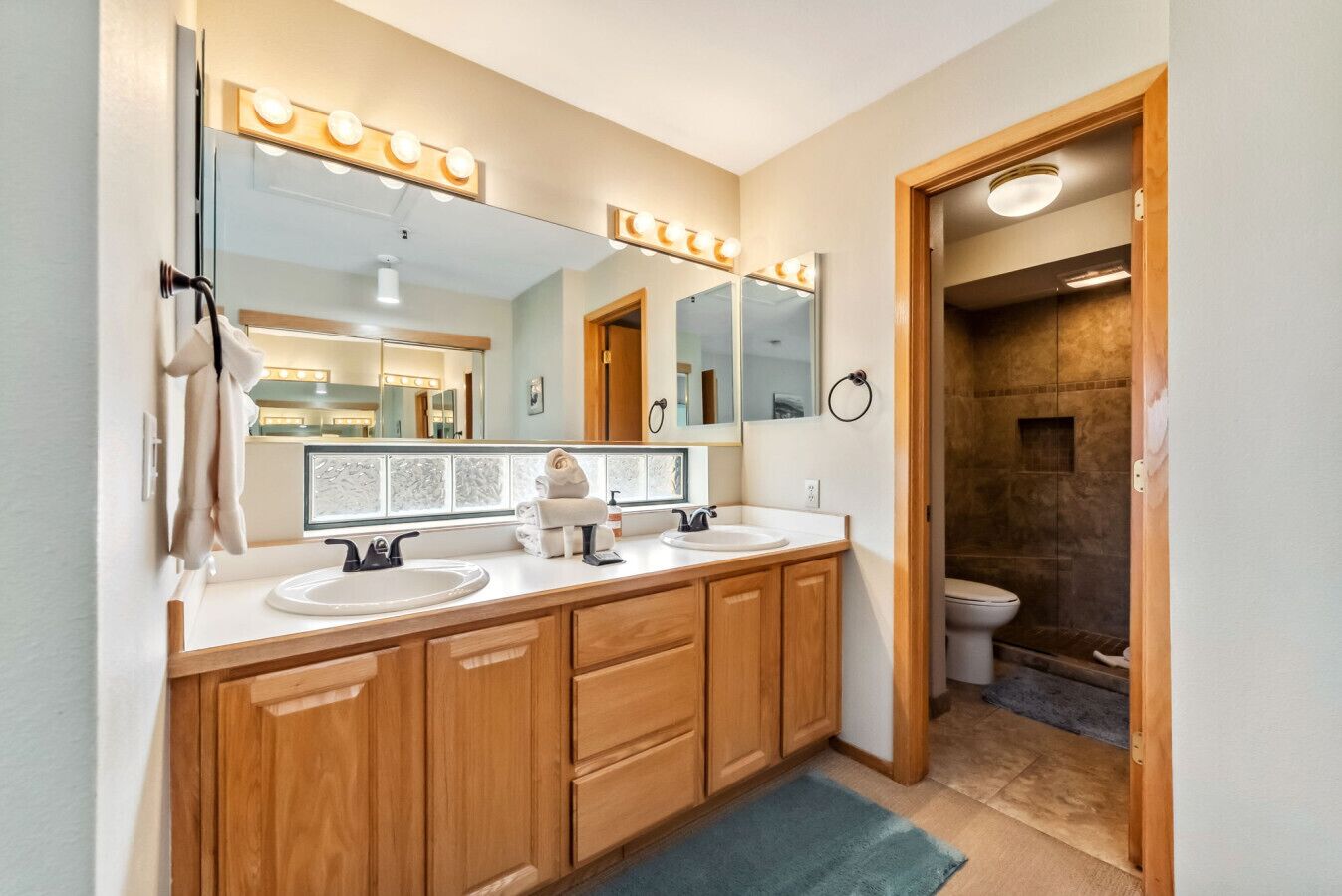 A bathroom with a vanity, two sinks, a large mirror with lights, a towel rack, and a shower area.