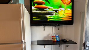 Uma televisão fixada na parede exibe uma cena da natureza. Abaixo da TV, há uma prateleira com duas garrafas e um controle remoto. Dois banquinhos de madeira estão dispostos em frente à prateleira.