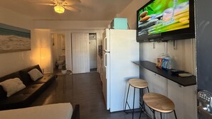 Um espaço de estar compacto com cozinha compacta, ventoinha no teto, TV acima do balcão e pequena área de estar com banquinhos.