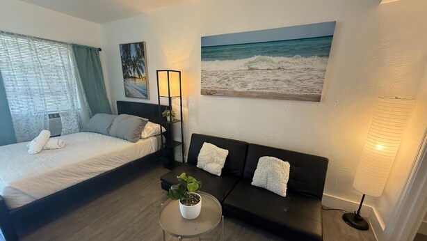 Quarto de hotel com uma cama, um sofá, uma mesinha com uma planta e um quadro de uma praia.