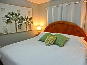 A neatly made bed with white linens and two green pillows, a wooden headboard, a nightstand with a lamp, and a framed picture of palm trees on the wall. - The Salty Mangrove | Dockside Retreat w/ Pool & Kayaks (Hernando Beach)