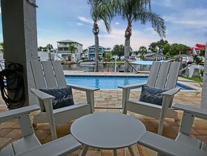 A poolside area with two white lounge chairs and a small table, a clear view of the pool, and a backdrop of buildings and palm trees. - The Salty Mangrove | Dockside Retreat w/ Pool & Kayaks (Hernando Beach)