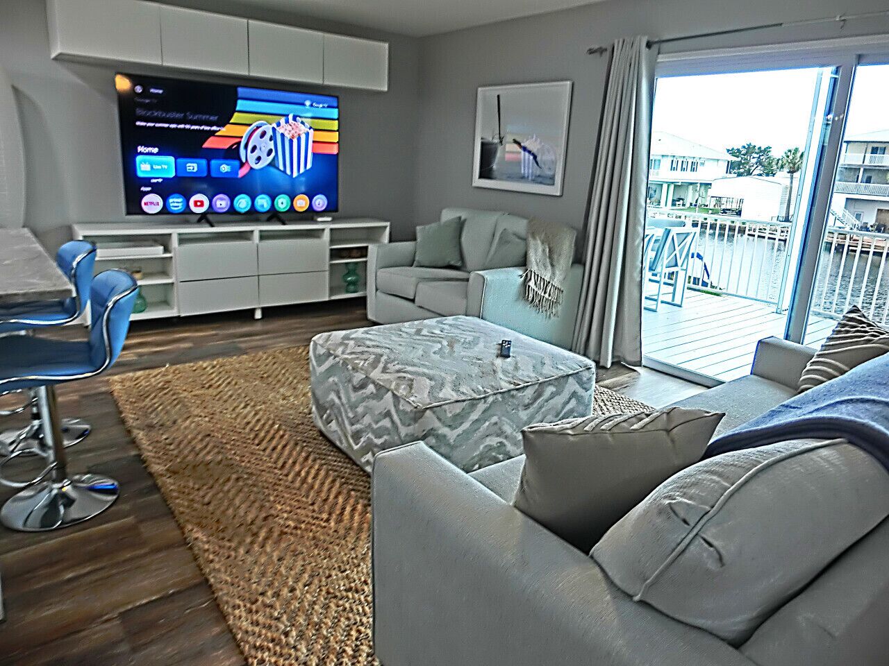A modern living room with a flat-screen TV, grey sofas, a patterned ottoman, a blue chair, and a view of a balcony with a railing.