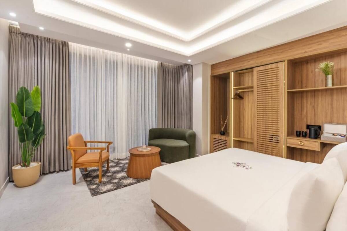 Una habitación de hotel moderna con una cama grande, un armario de madera, un sillón verde, una pequeña mesa redonda y una planta en maceta.
