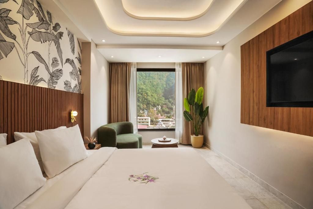 A modern hotel room with a large bed, a green armchair, a small table, a potted plant, and a TV mounted on the wall.