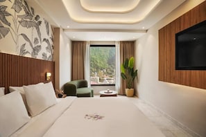 A modern hotel room with a large bed, a green armchair, a small table, a potted plant, and a TV mounted on the wall. - Zana By The Ganges, Rishikesh (Narendranagar)