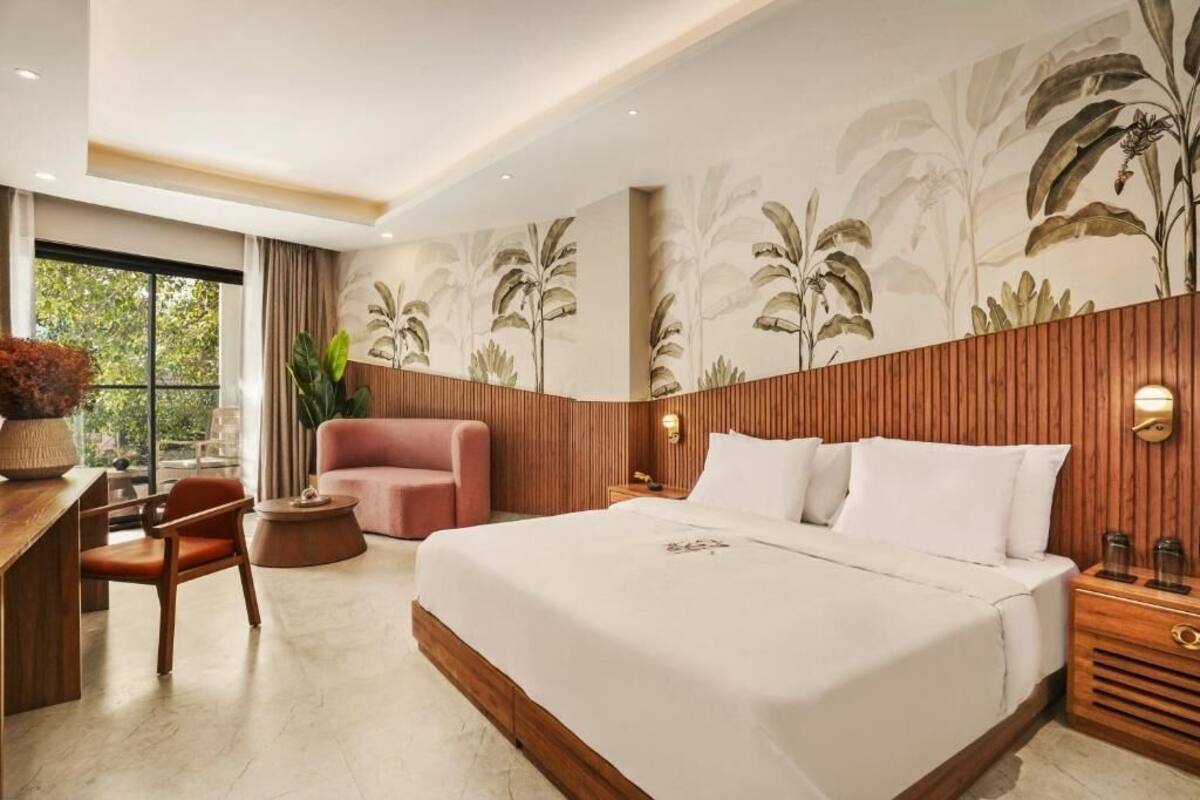 Una habitación de hotel moderna con una cama grande, cabecera de madera, un escritorio con silla, un sillón rosa y un ventanal con vistas a zonas verdes.
