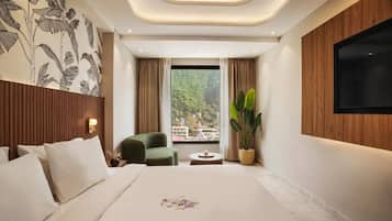 A modern hotel room with a large bed, a green armchair, a small table, a potted plant, and a TV mounted on the wall.
