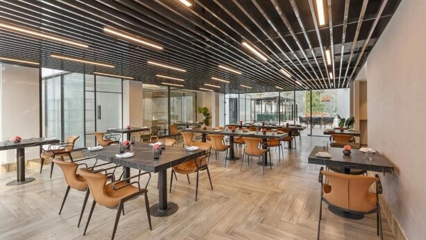 A modern dining area with tables and chairs, large windows, and a ceiling with linear lighting fixtures. - Zana By The Ganges, Rishikesh (Narendranagar)