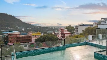 A swimming pool with a tiled border, surrounded by buildings and a hilly landscape in the background.