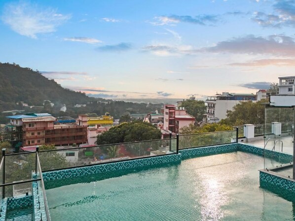 A swimming pool with a tiled border, surrounded by buildings and a hilly landscape in the background. - Zana By The Ganges, Rishikesh (Narendranagar)
