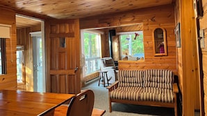 A wooden cabin interior with a dining table, a striped sofa, and a window with a view of greenery. - Peaceful Waterfront Cottage on Ottawa River – Nature Lover’s Retreat! (Westmeath)