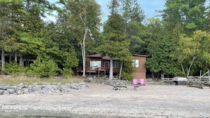 A cabin with a deck, surrounded by trees and a gravel area. - Peaceful Waterfront Cottage on Ottawa River – Nature Lover’s Retreat! (Westmeath)