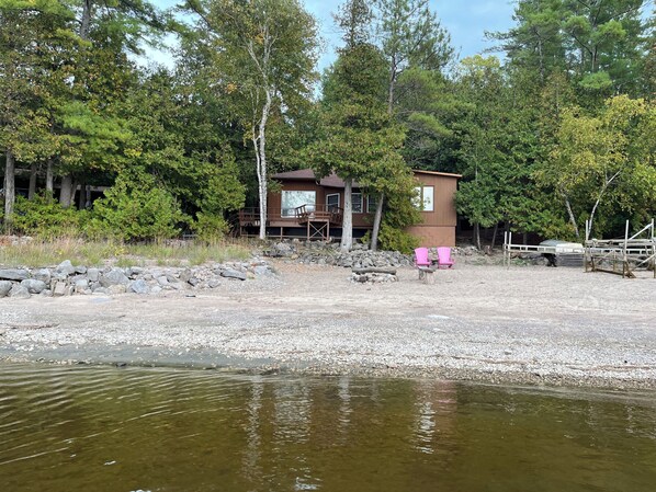 A cabin with a deck, surrounded by trees and a gravel area.