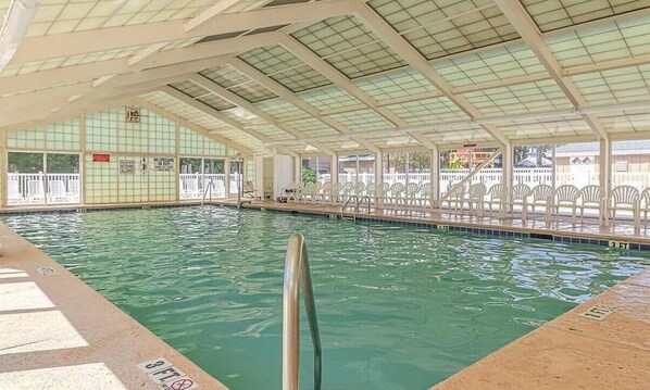 An indoor swimming pool with clear water, a diving board, and a covered seating area with chairs.