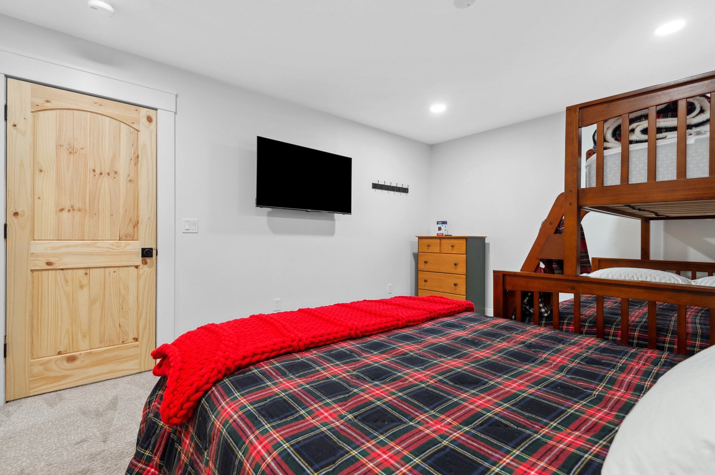Een slaapkamer met een houten stapelbed, een aan de muur bevestigde televisie, een rode deken op het bed en een houten deur.