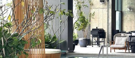 A modern outdoor pool area with a wooden seating area, a grill, and potted plants.