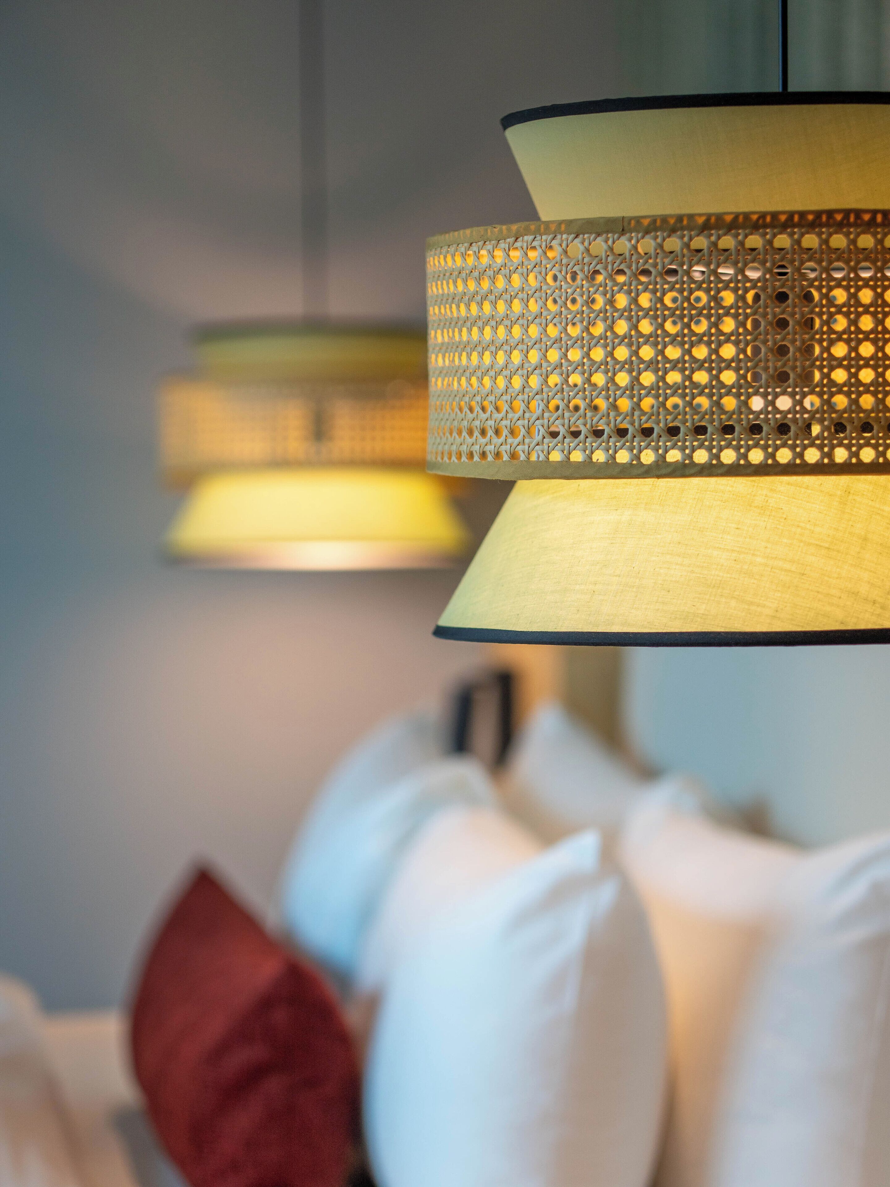 A close-up of a hanging lamp with a perforated shade, a bed with white and red pillows, and a blurred background suggesting a room setting.