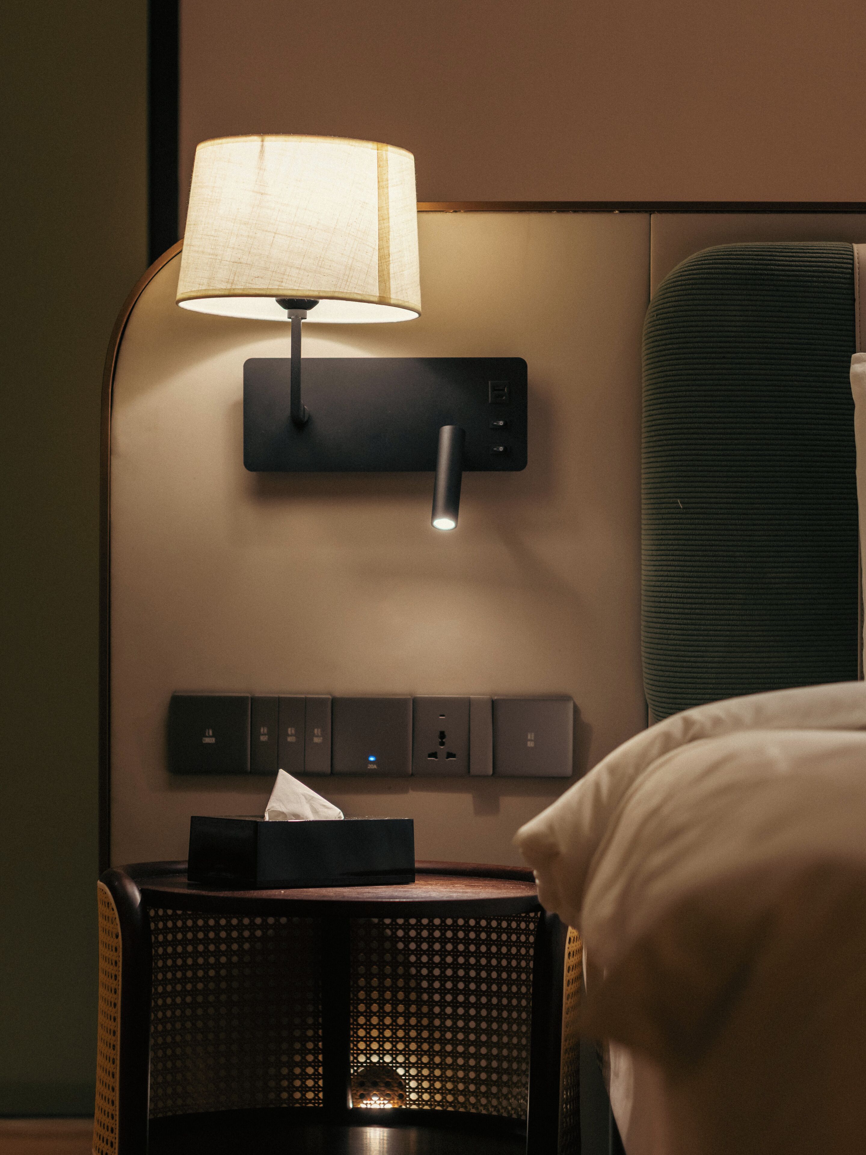A hotel room with a bedside table, a lamp, a power strip, and a green upholstered headboard.