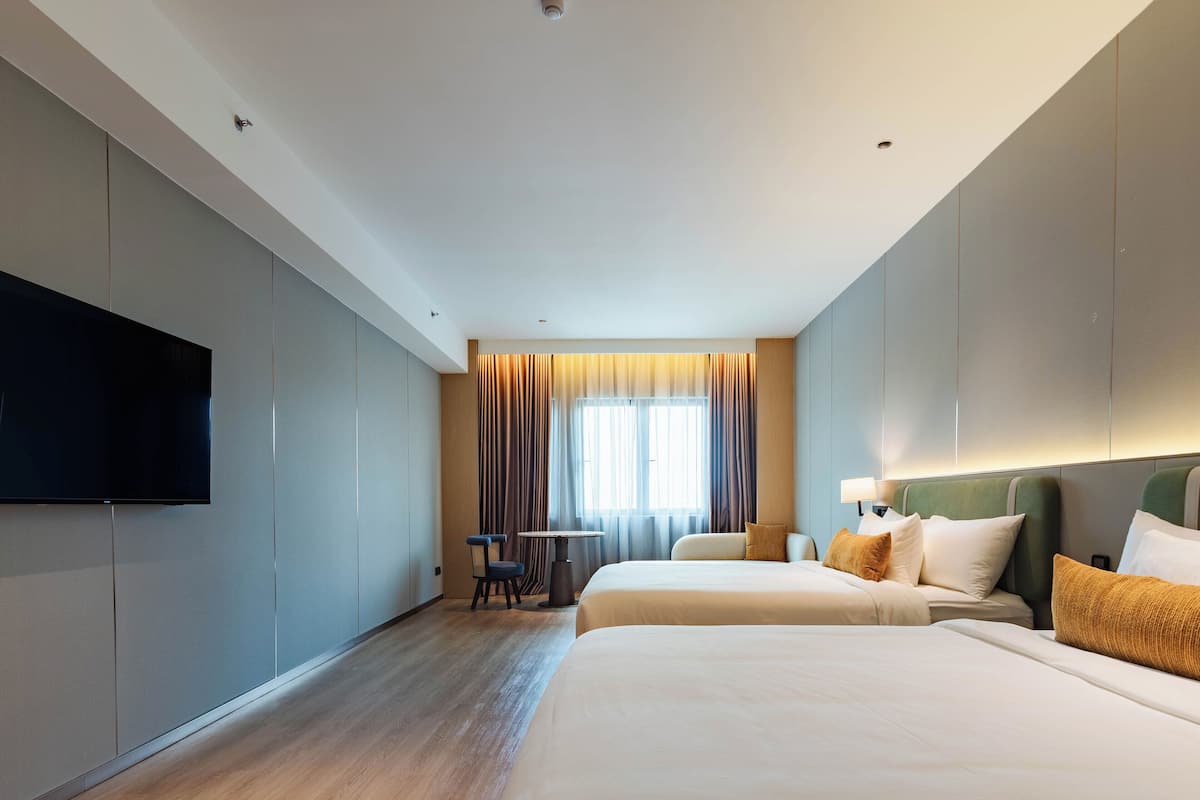 Une chambre d’hôtel avec deux lits, un téléviseur à écran plat fixé au mur, une petite table avec des chaises et une fenêtre avec des rideaux.