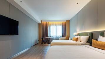 A hotel room with two beds, a flat-screen TV mounted on the wall, a small table with chairs, and a window with curtains.