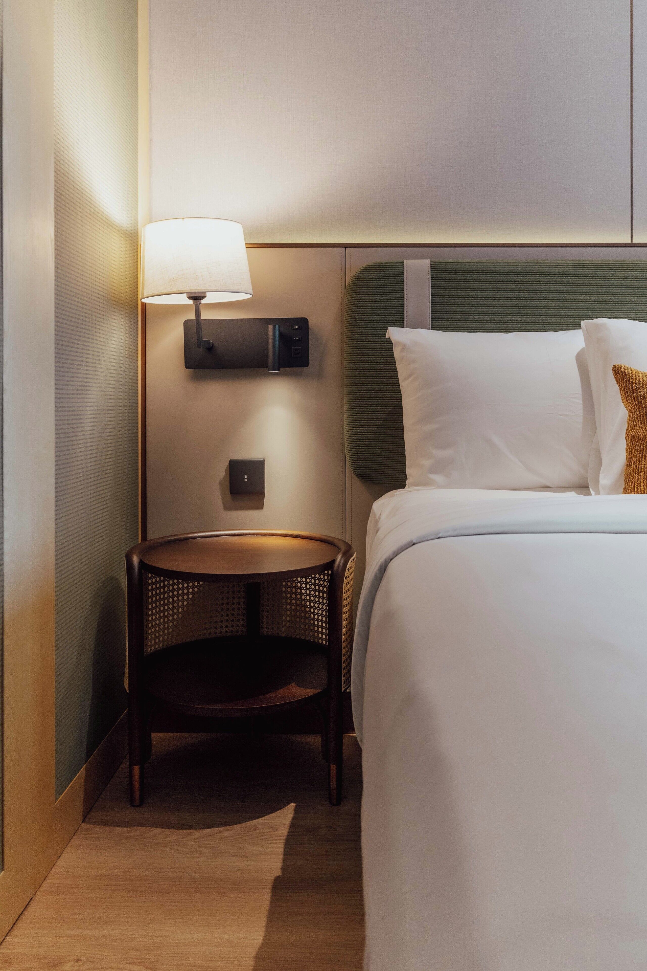A neatly arranged hotel room with a bed, a bedside table with a lamp, and a wall-mounted control panel.