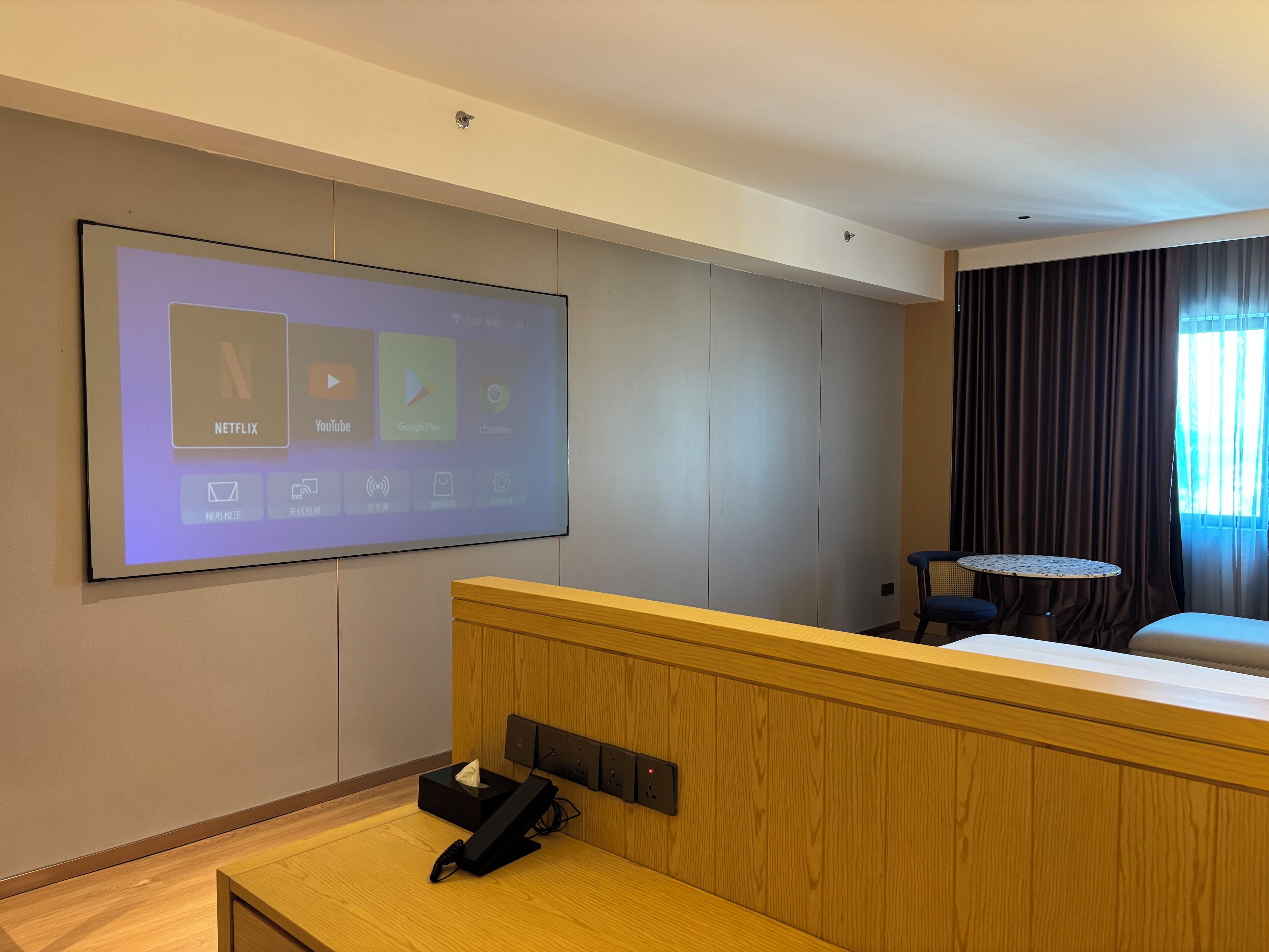 Una camera d'albergo con un grande schermo che mostra applicazioni di streaming, una scrivania in legno con un telefono, un tavolino e un letto.