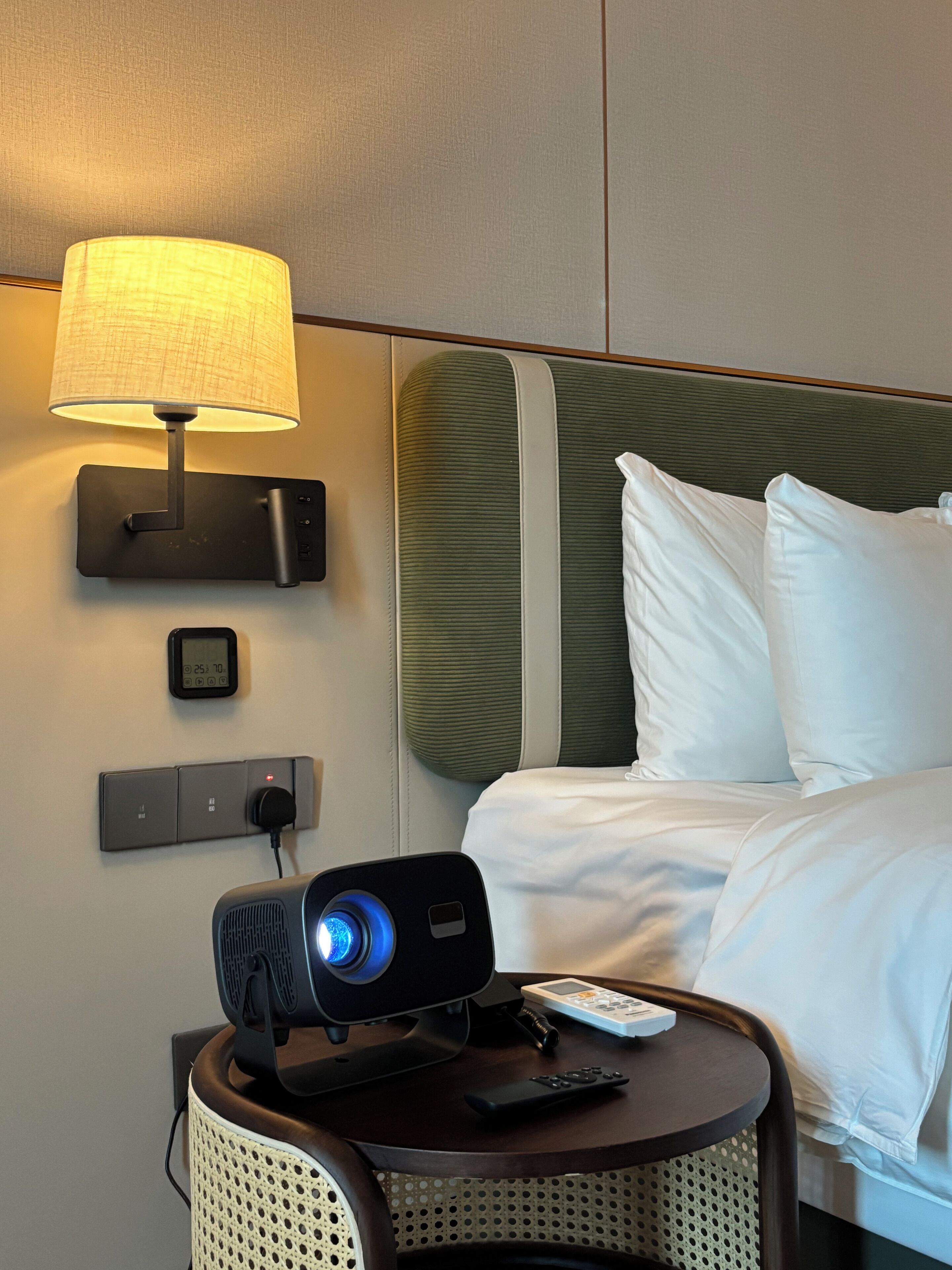 Habitación de hotel con cama, mesita de noche con lámpara, control remoto y un proyector.