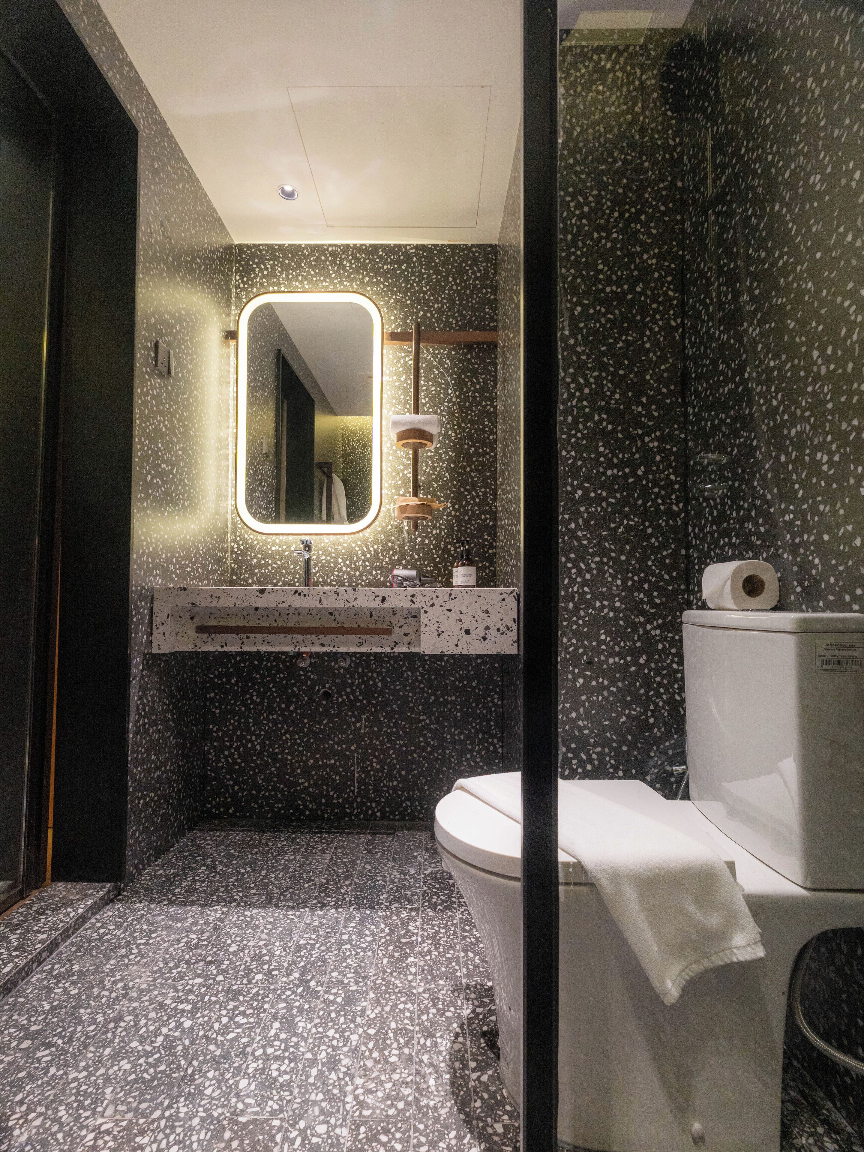 A modern bathroom with a speckled floor, a mirror with a light, a toilet, and a sink with a towel hanging on it.
