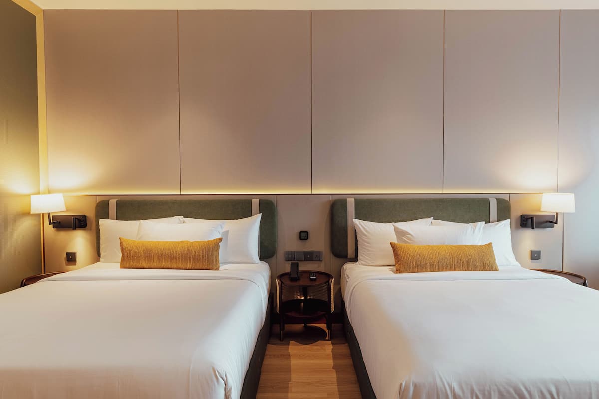 Une chambre d’hôtel avec deux lits, chacun recouvert de draps blancs et agrémenté d’un seul oreiller jaune. Il y a deux tables de chevet avec des lampes, et une petite table d’appoint entre les lits.