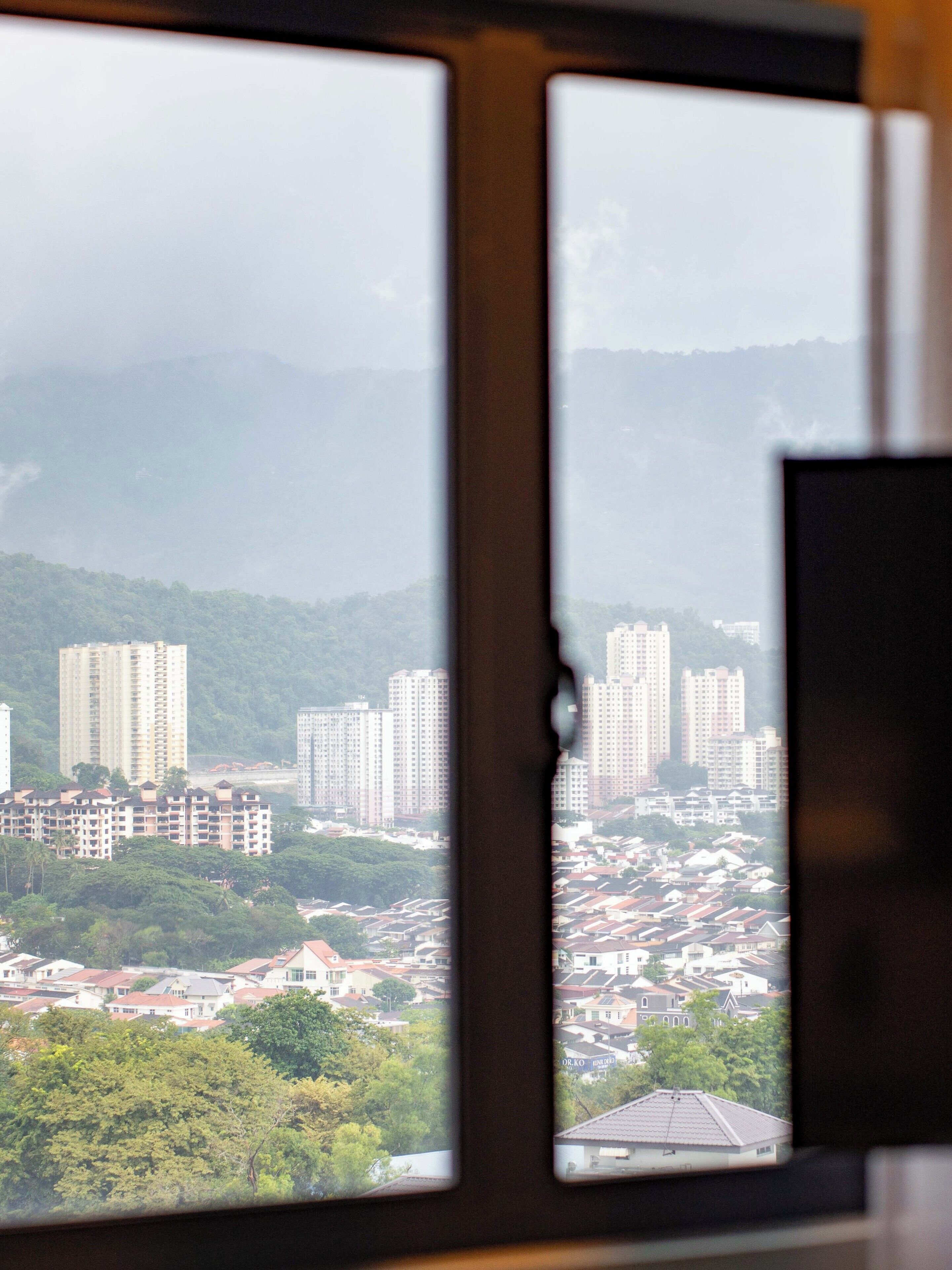 Visão pela janela com borda rasgada, mostrando um horizonte urbano com vários edifícios altos, uma área residencial densa e montanhas ao fundo.