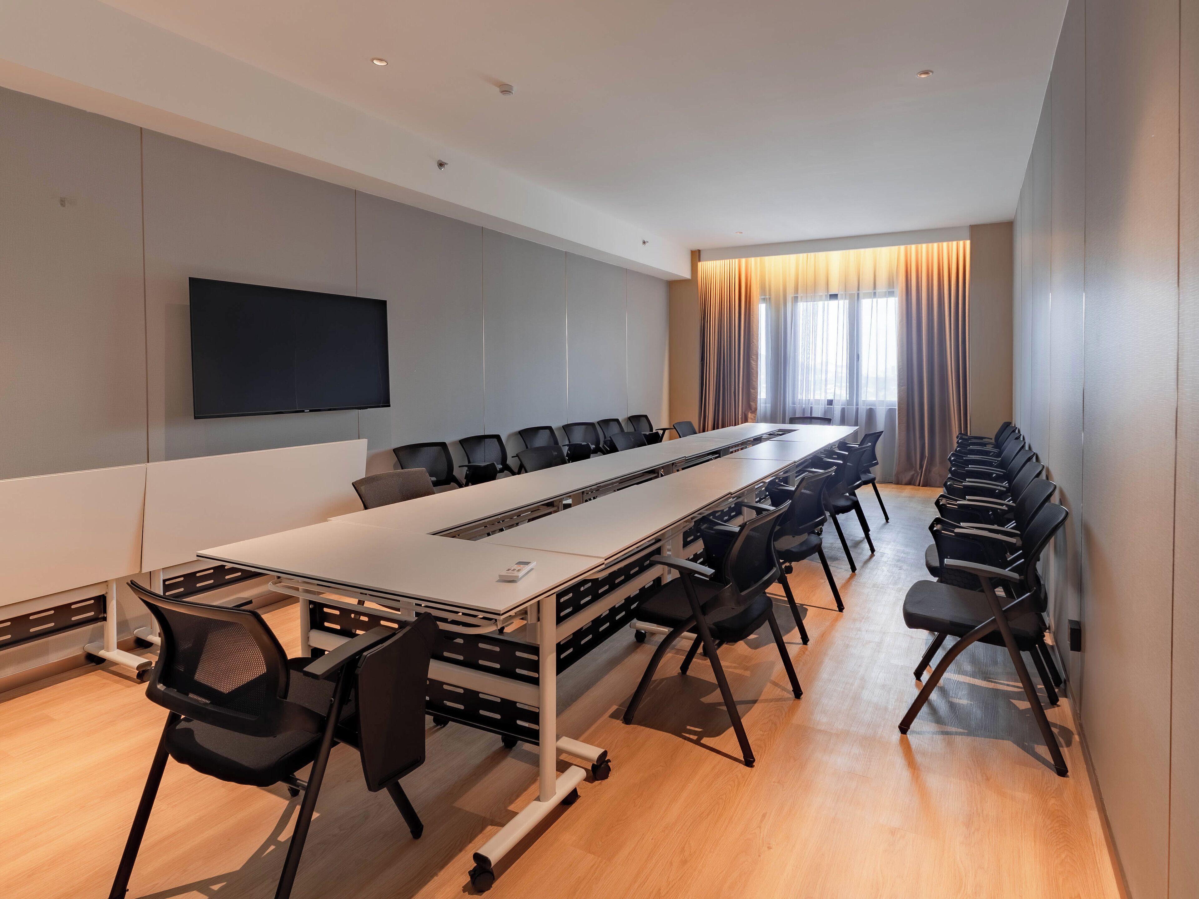A conference room with a long table, chairs, a flat-screen TV, and a large window with curtains.