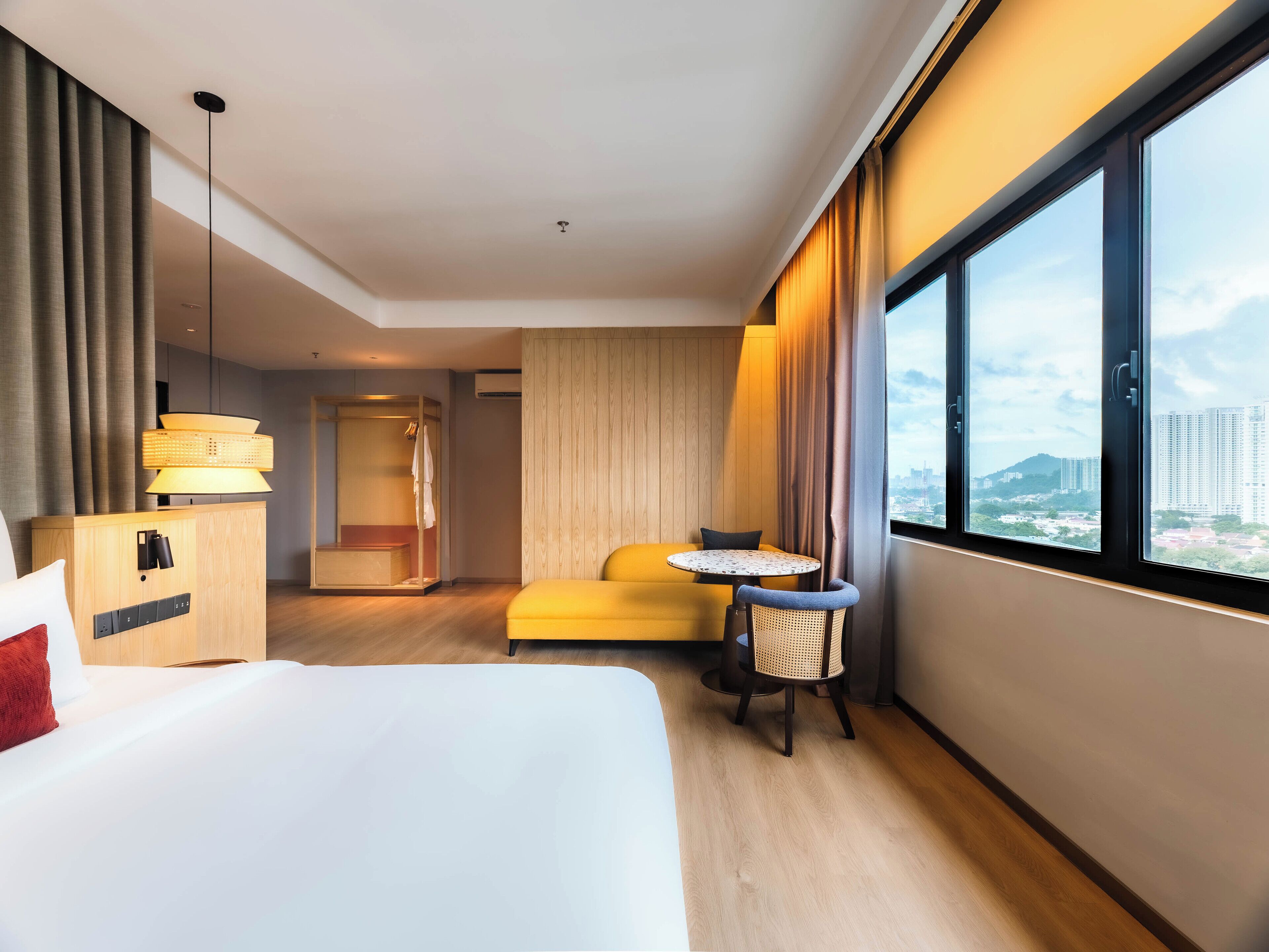 Habitación de hotel con cama, una mesita con silla, armario, escritorio y una ventana grande con vista a la ciudad.