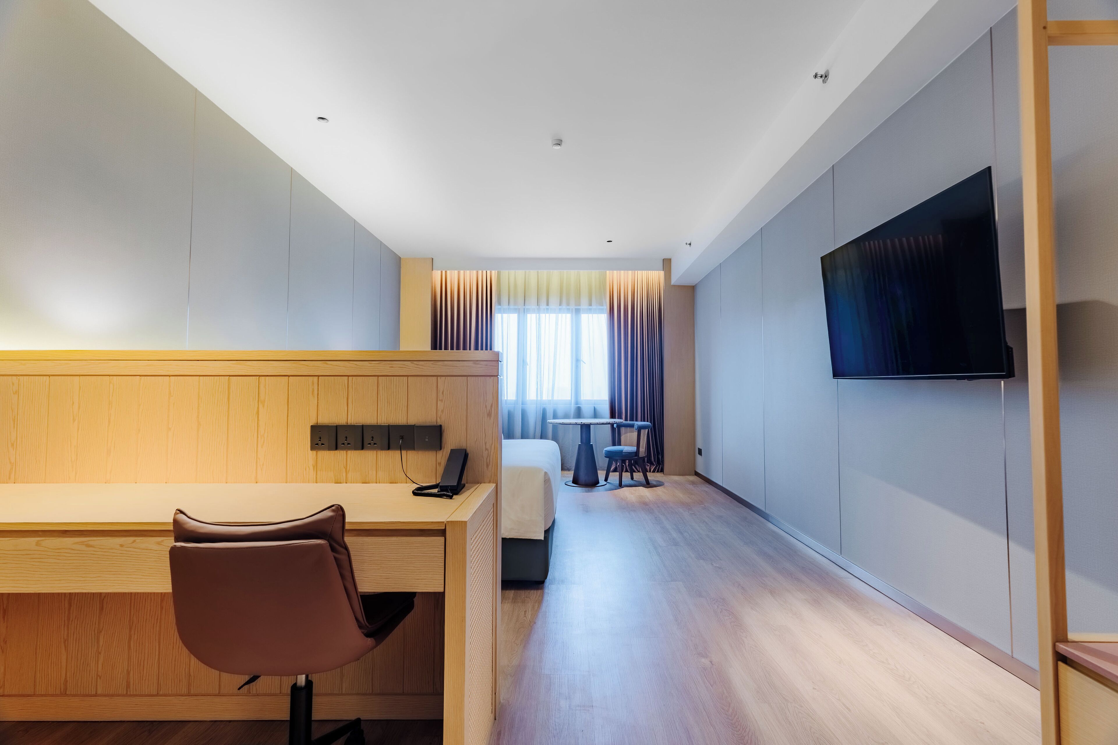 A hotel room with a wooden desk, a bed, a TV, and a window with curtains.