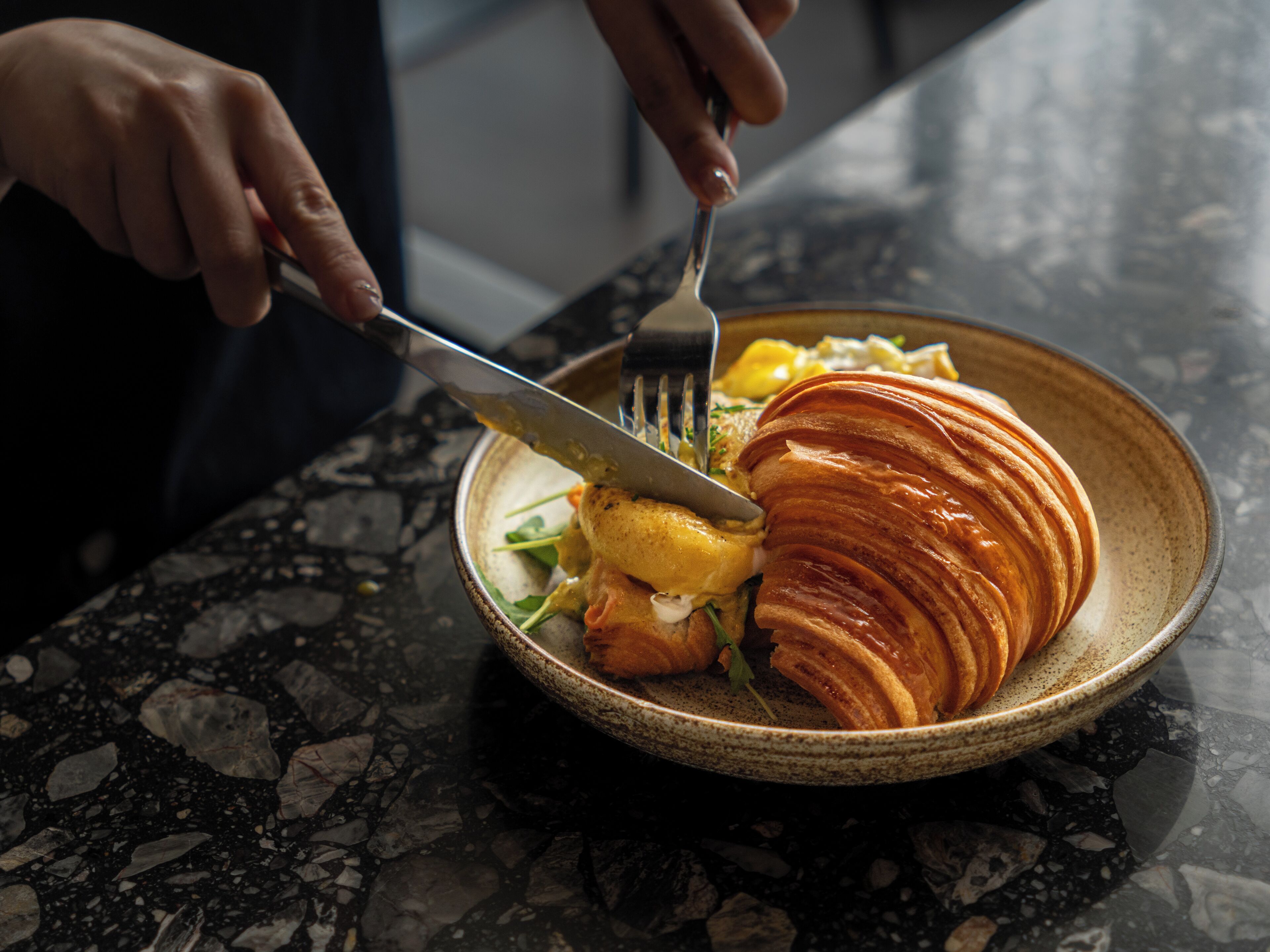 Une personne coupe un croissant sur une assiette en céramique à l’aide d’une fourchette et d’un couteau.