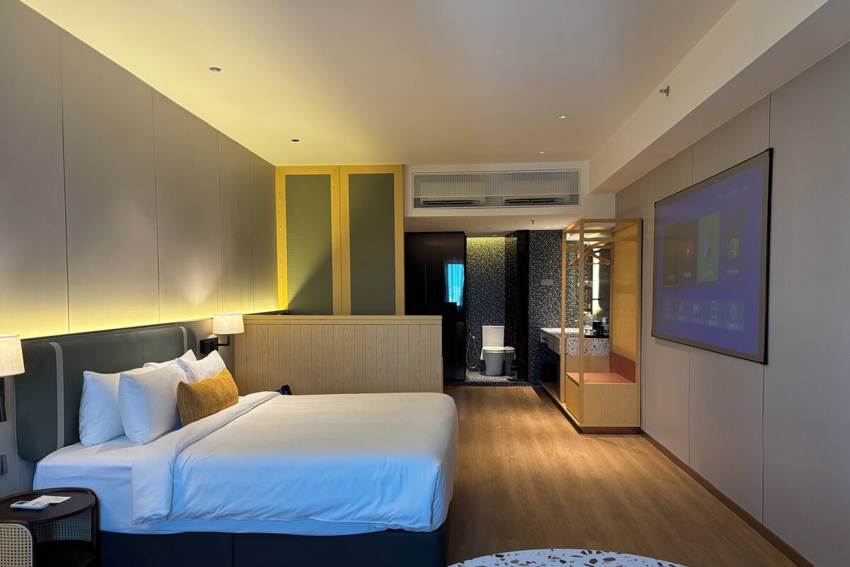 Une chambre d’hôtel moderne avec un grand lit, une table de chevet, une table ronde et une salle de bain visible à travers une porte ouverte.