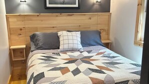 Une chambre à coucher avec une tête de lit en bois, une couvre-lit matelassé, deux lampes de chevet, un tableau encadré au mur et une fenêtre sur le côté droit.