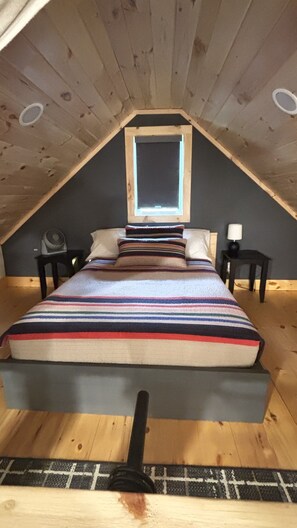 Une chambre à coucher aménagée dans un grenier, avec un plafond en pente, un lit recouvert d’une couverture à rayures, une table de chevet avec une lampe et une fenêtre offrant une vue.